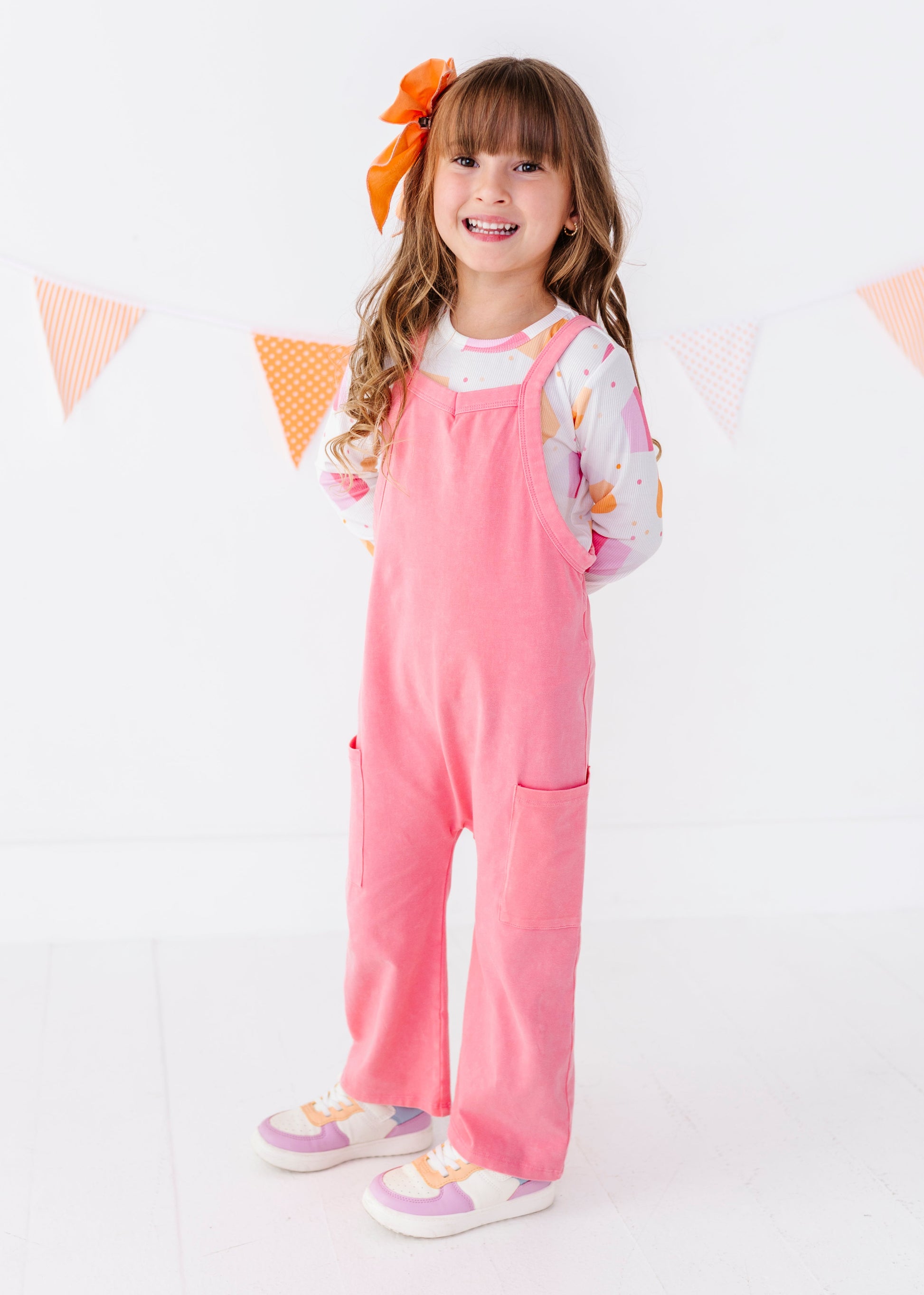 A young girl with a brown hair bow stands wearing a pink corduroy overall jumpsuit over a white long-sleeve floral shirt and pastel sneakers. She is posing in front of a white wall with a decorative orange and white bunting banner.