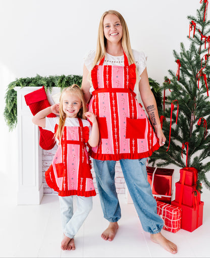 A woman and a young girl wearing matching red and pink quilted aprons pose in front of a white fireplace mantel decorated with greenery and Christmas stockings, with a Christmas tree and wrapped presents to the right.