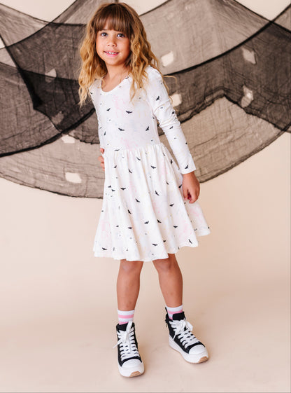 Young girl wearing a white dress with black patterns, standing against a neutral background.