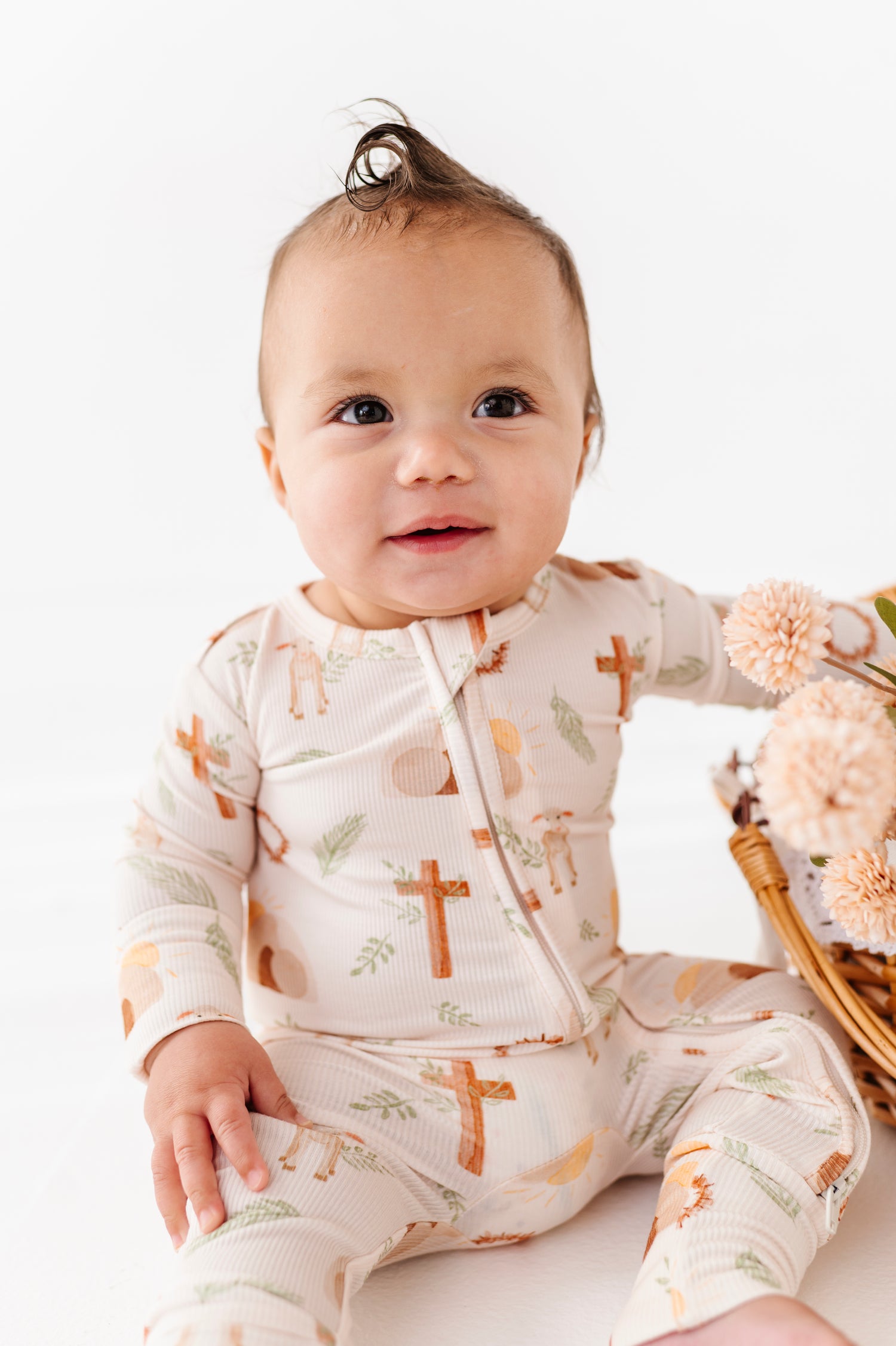 Baby wearing a patterned onesie sitting on a white surface with a basket of flowers in the background.