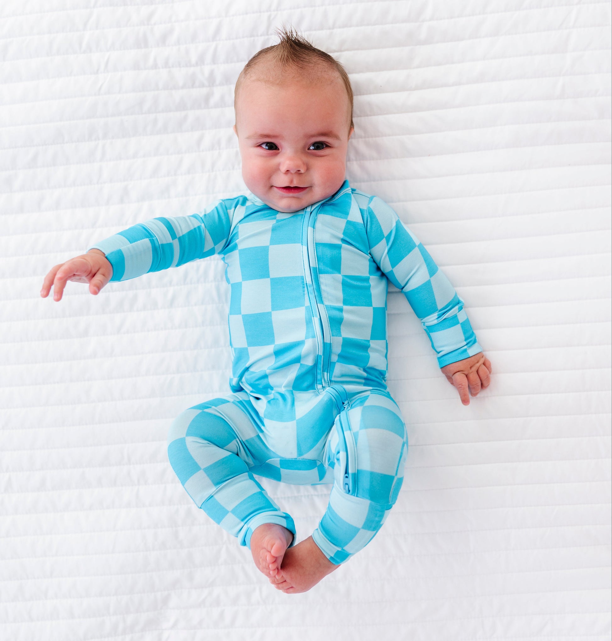 A baby lying on a white quilted blanket, wearing a light blue and white checkered long-sleeve footed sleeper.