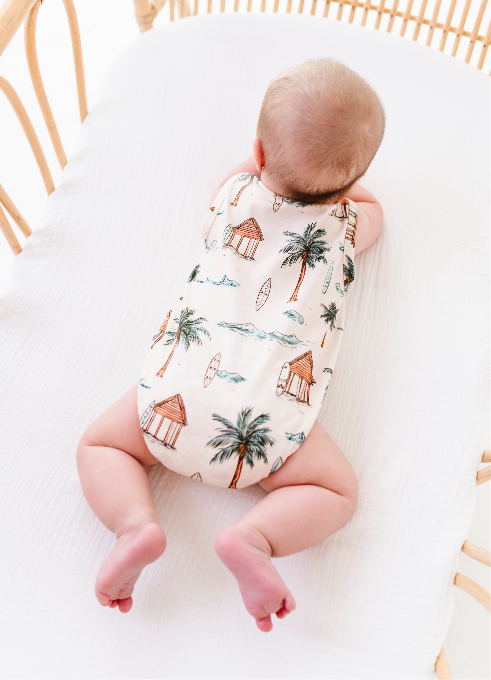 Baby in a patterned onesie lying in a crib with white bedding and wooden crib bars.
