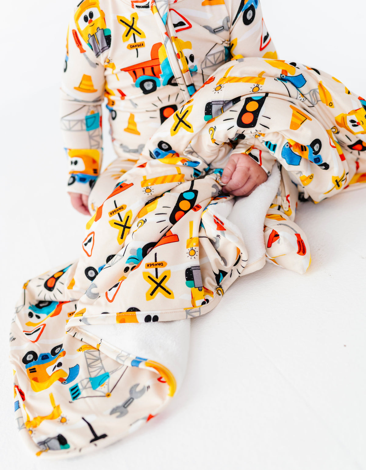A baby wearing a cream-colored one-piece jumpsuit covered in a colorful cartoon construction worker pattern, sitting on a white floor and wrapping a matching blanket around themselves.