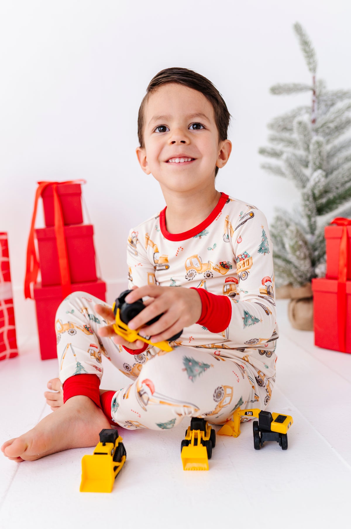 A young boy sitting on a white floor wearing a festive Christmas-themed pajama set with construction vehicles, holding a toy construction vehicle. Red gift boxes and a decorated Christmas tree are visible in the background.