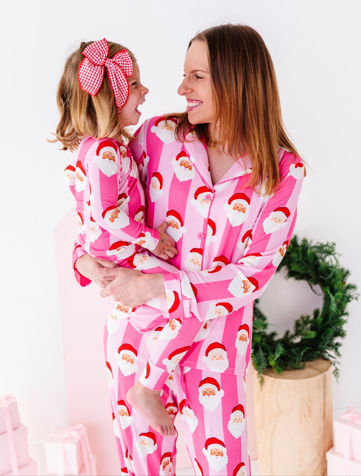 A woman and a young girl wearing matching pink Santa bamboo pajamas with a white striped background and a Santa face print. The woman holds the girl who has a pink gingham bow in her hair.