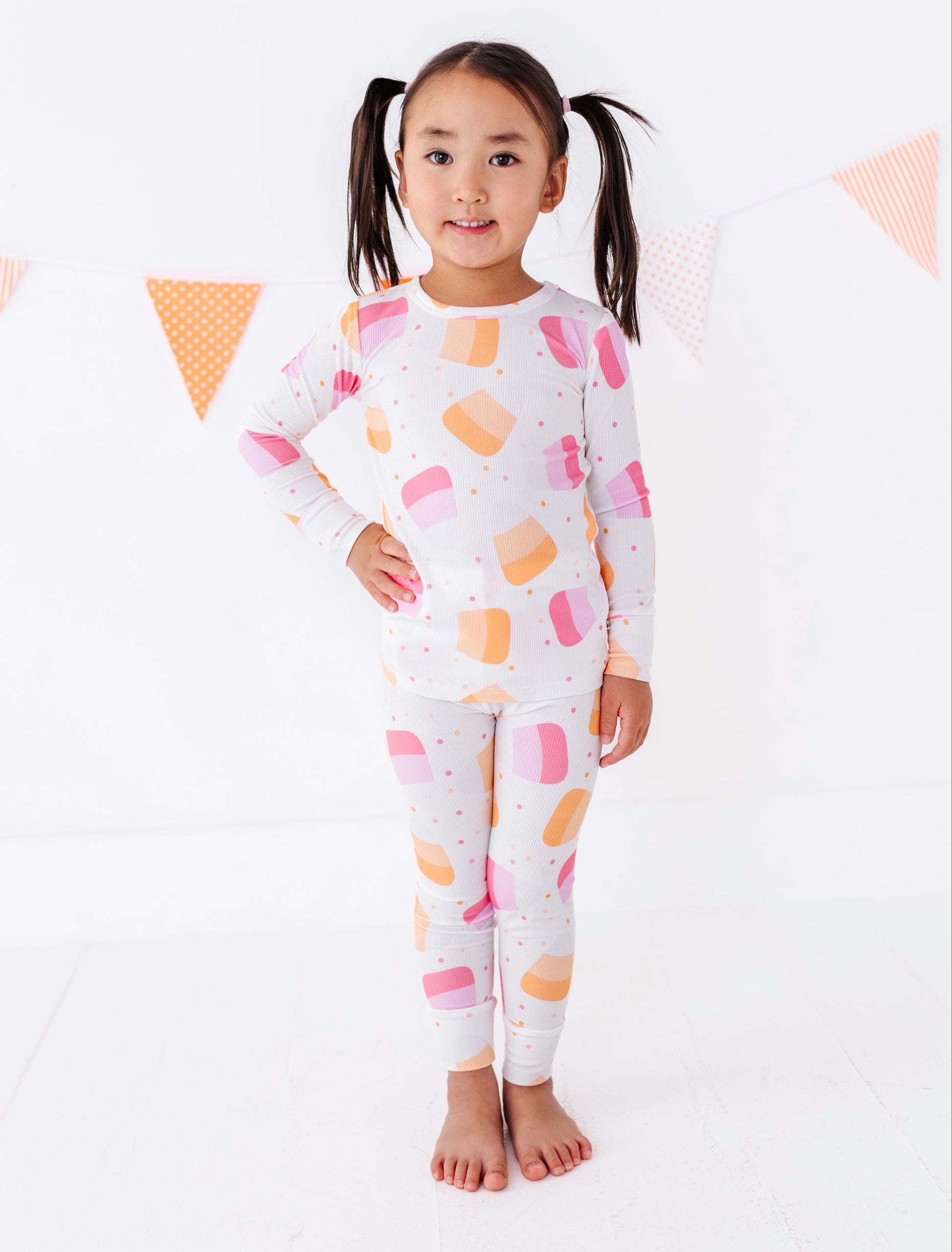 A young girl with dark hair in pigtails stands barefoot in a white studio setting, wearing a matching long-sleeve top and leggings set with a colorful ice cream cone pattern in pink, orange, and yellow on a white background.