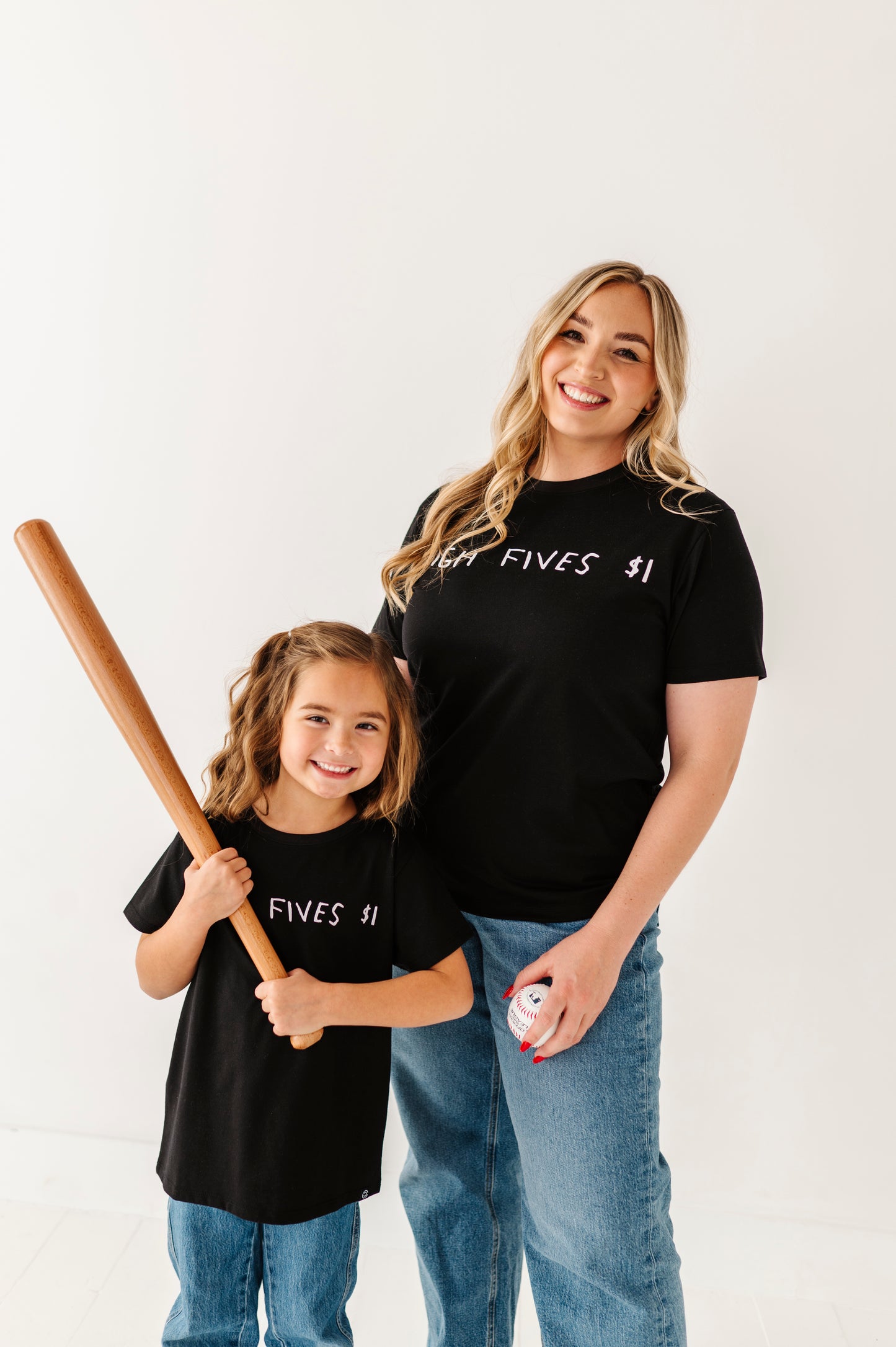 Woman and child wearing matching black t-shirts with text, holding a baseball bat and ball.