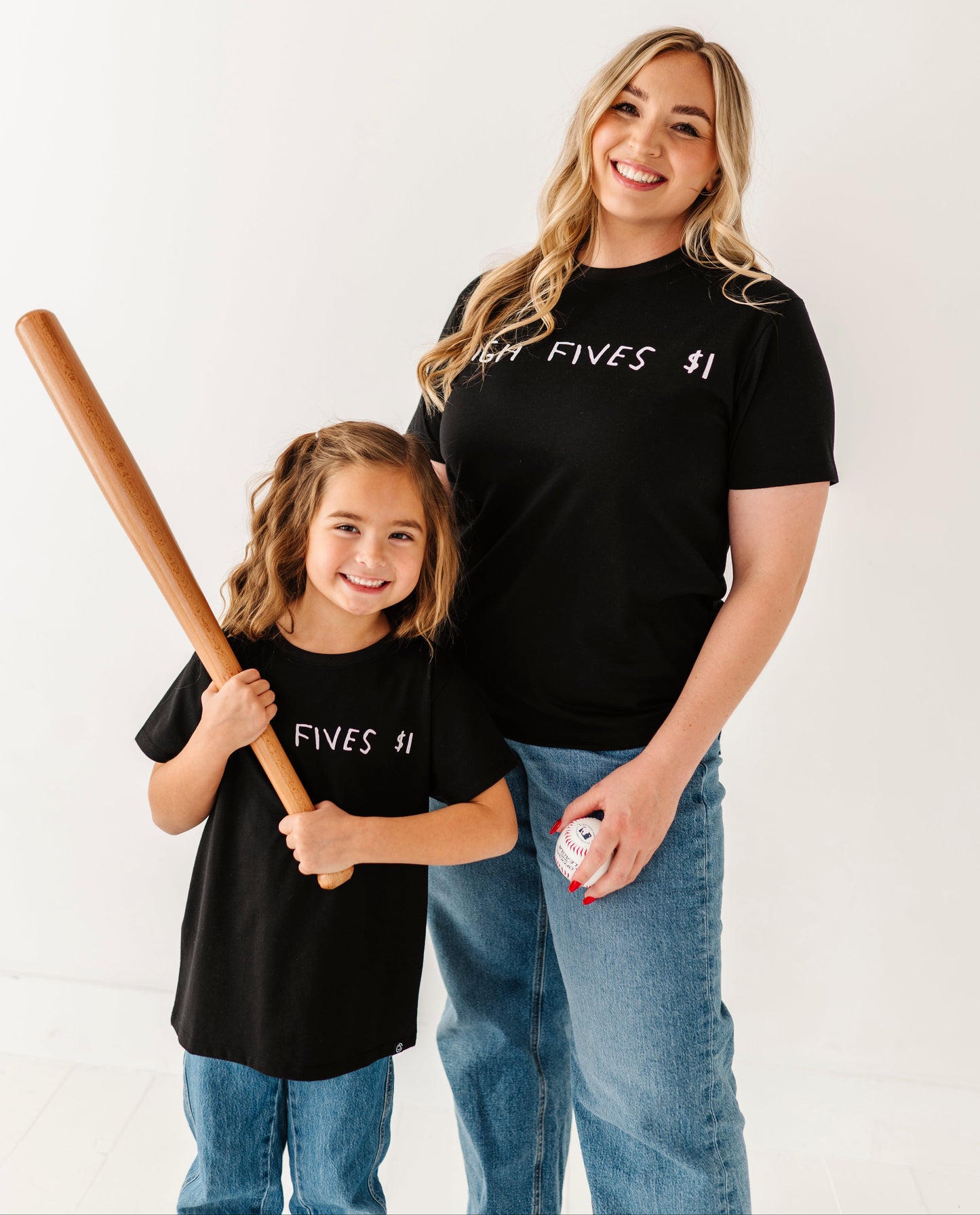 Woman and child wearing matching black t-shirts with text, holding a baseball bat and ball.