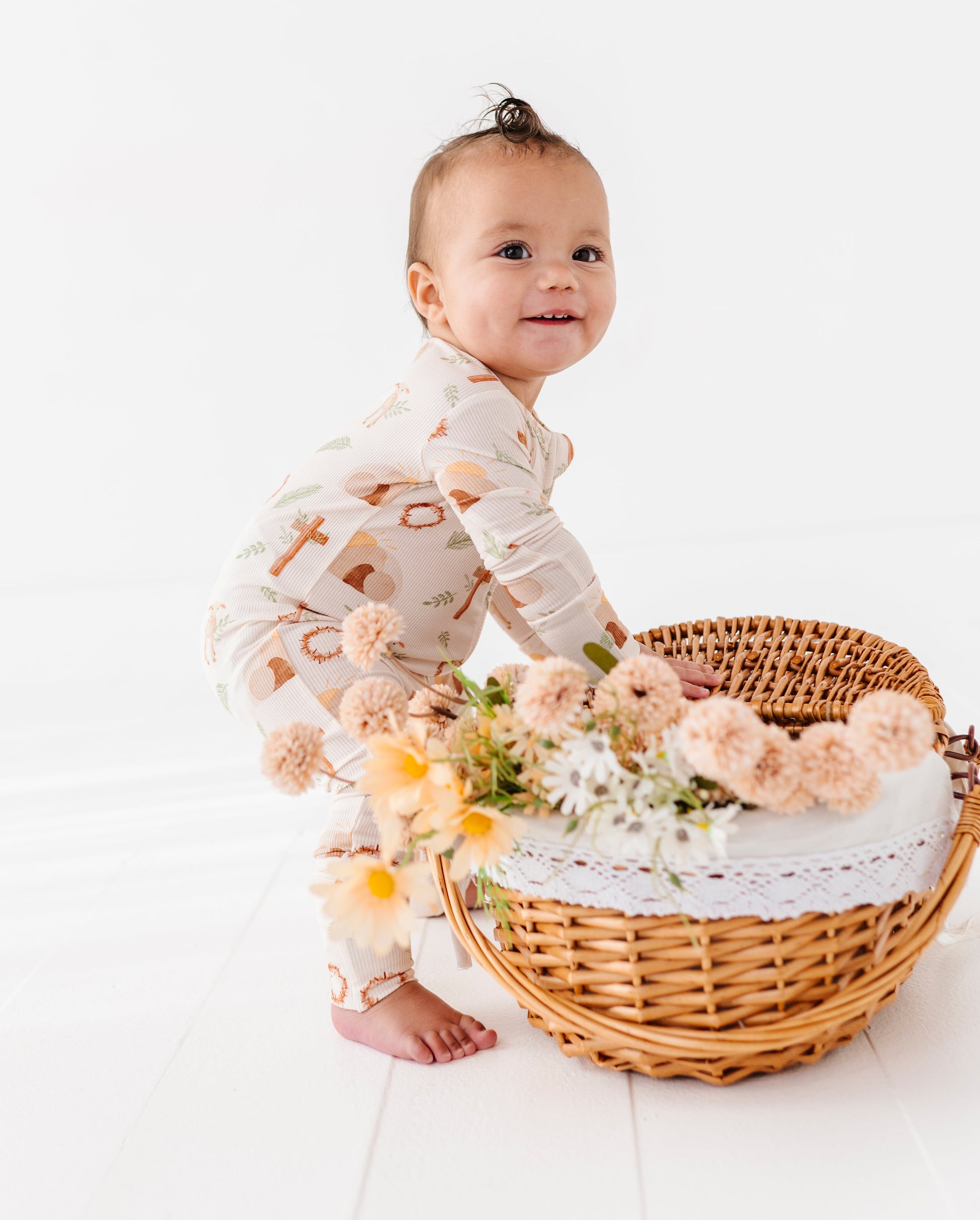Baby in a cross patterned Easter romper standing next to a wicker basket with flowers on a white background