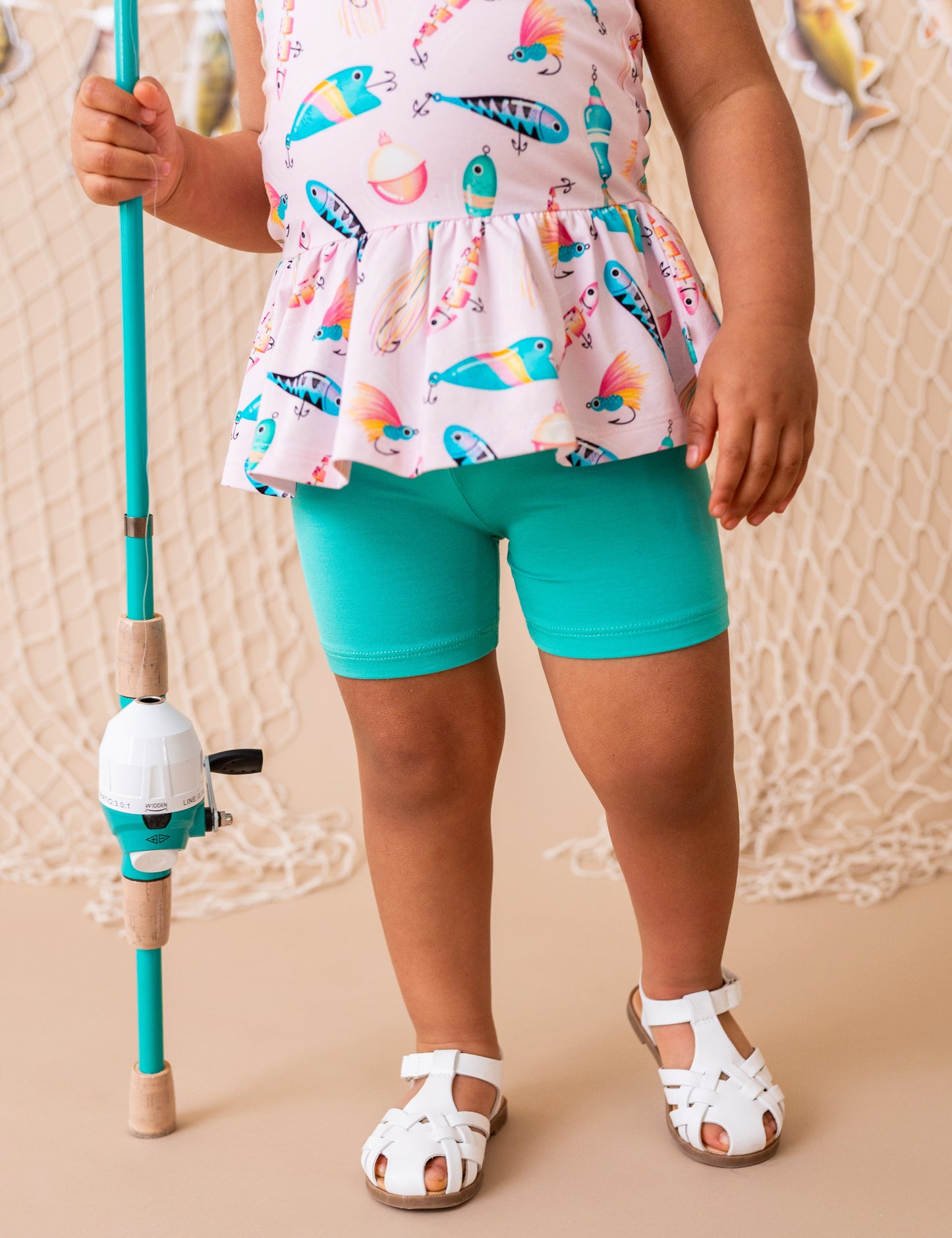 Child wearing a colorful top and teal shorts holding a fishing rod on a beige background