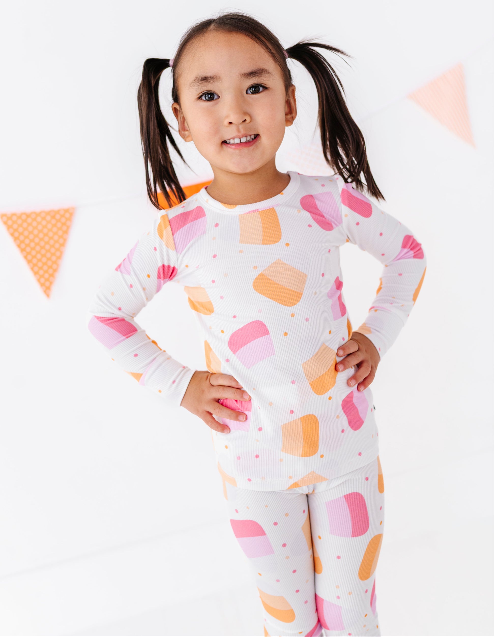 A young girl with dark hair in pigtails stands smiling, wearing a white long-sleeve pajama set with a colorful pattern of pink, orange, and yellow ice cream scoops and small pink dots. She is posed against a clean white background with subtle orange and pink triangular bunting decorations.
