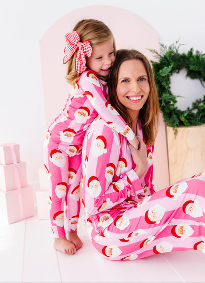 A woman and a young girl wearing matching pink Santa-themed bamboo pajamas, sitting together in a bright, festive room with a pink arch backdrop and a green wreath.