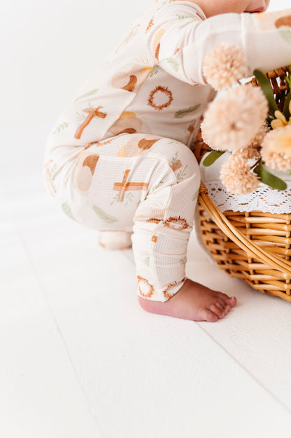 Baby wearing an Easter romper with crosses and religious elements, sitting next to a wicker basket filled with flowers on a white surface.
