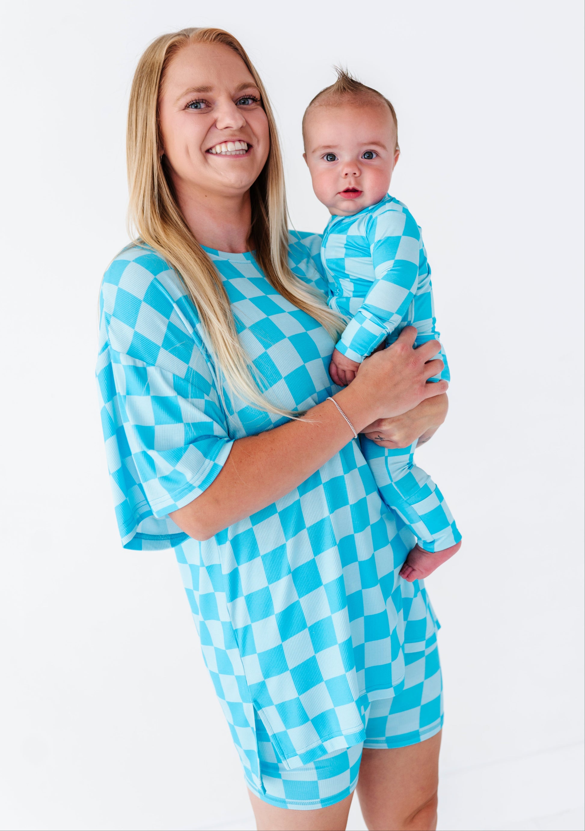 A smiling blonde woman holds a baby wearing a matching light blue and white checkered romper. The woman is wearing a matching oversized checkered top.