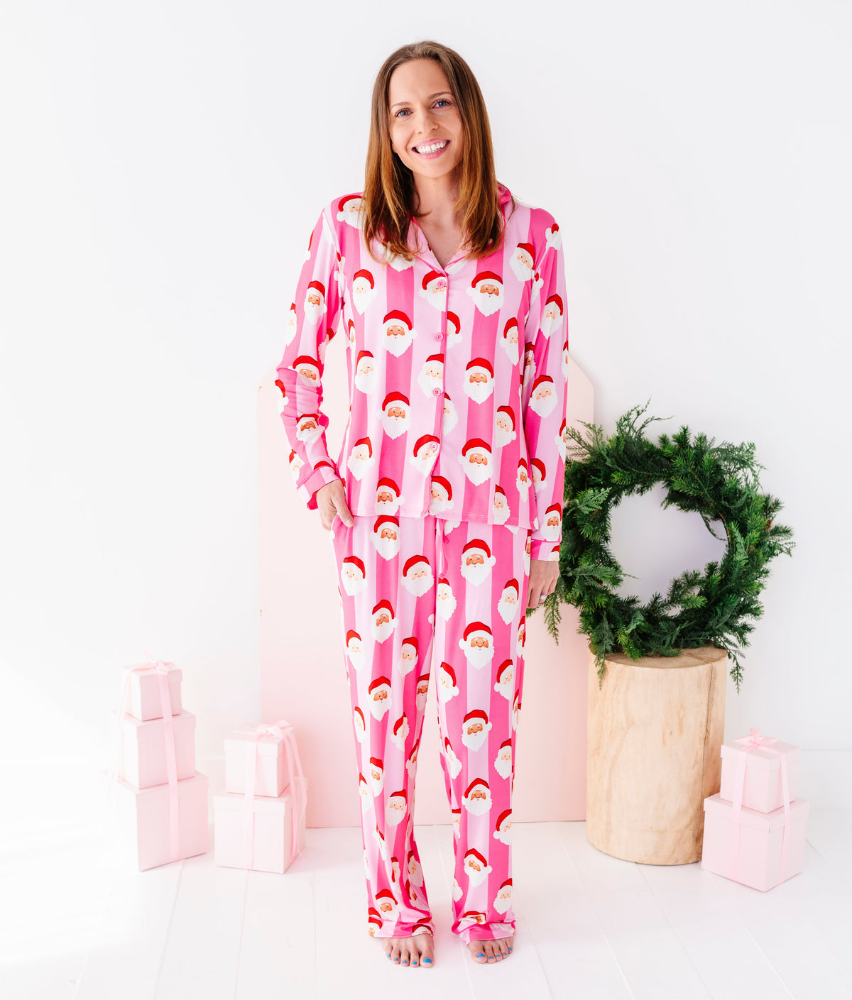 A woman wearing pink Santa Claus bamboo pajamas, standing in a bright white room with a green wreath and wrapped gifts in the background.