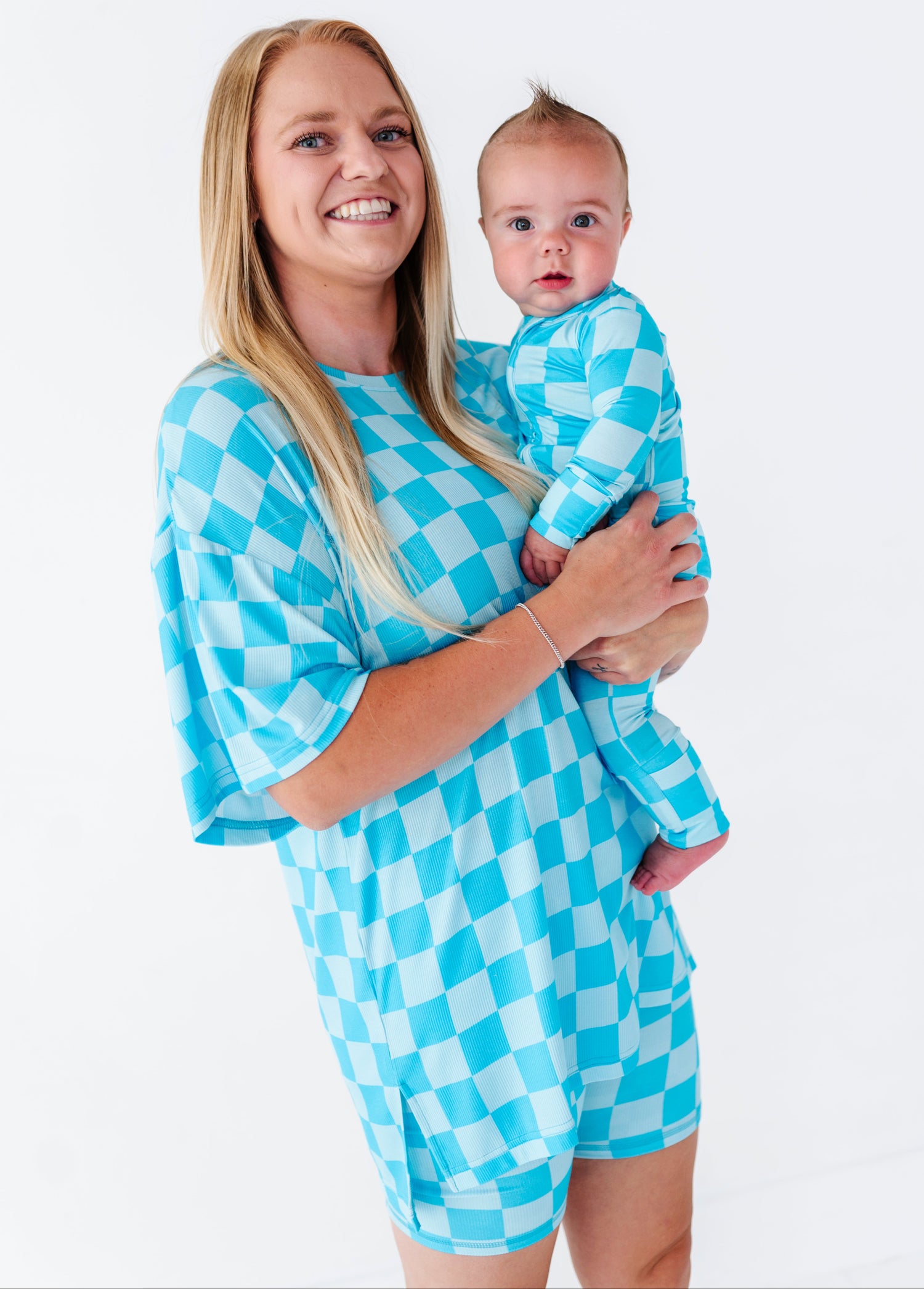 A smiling blonde woman holds a baby wearing a matching light blue and white checkered romper. The woman is wearing a matching oversized checkered top.