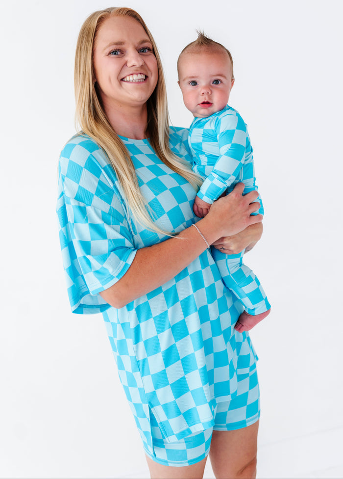 A smiling blonde woman holds a baby wearing a matching light blue and white checkered romper. The woman is wearing a matching oversized checkered top.