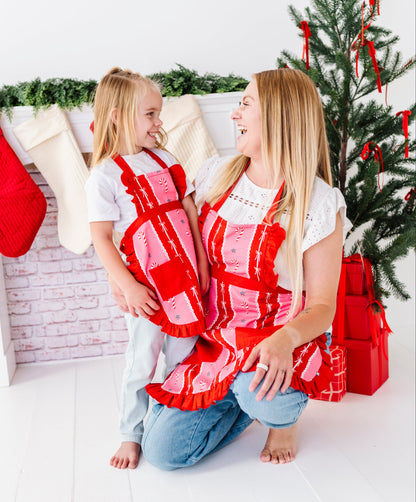 A woman and a young girl kneeling on a white floor in a decorated holiday room, both wearing matching red and pink patchwork aprons. A Christmas tree, stockings, and wrapped presents are visible in the background.