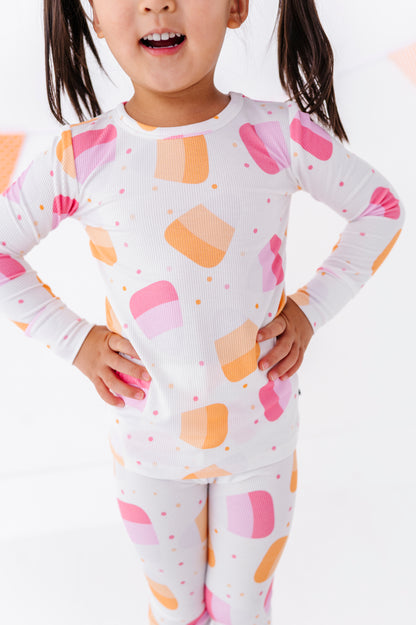 A young girl wearing a white long-sleeve top and matching leggings with a colorful abstract print of pink, orange, and peach shapes, standing with hands on hips against a bright white background.
