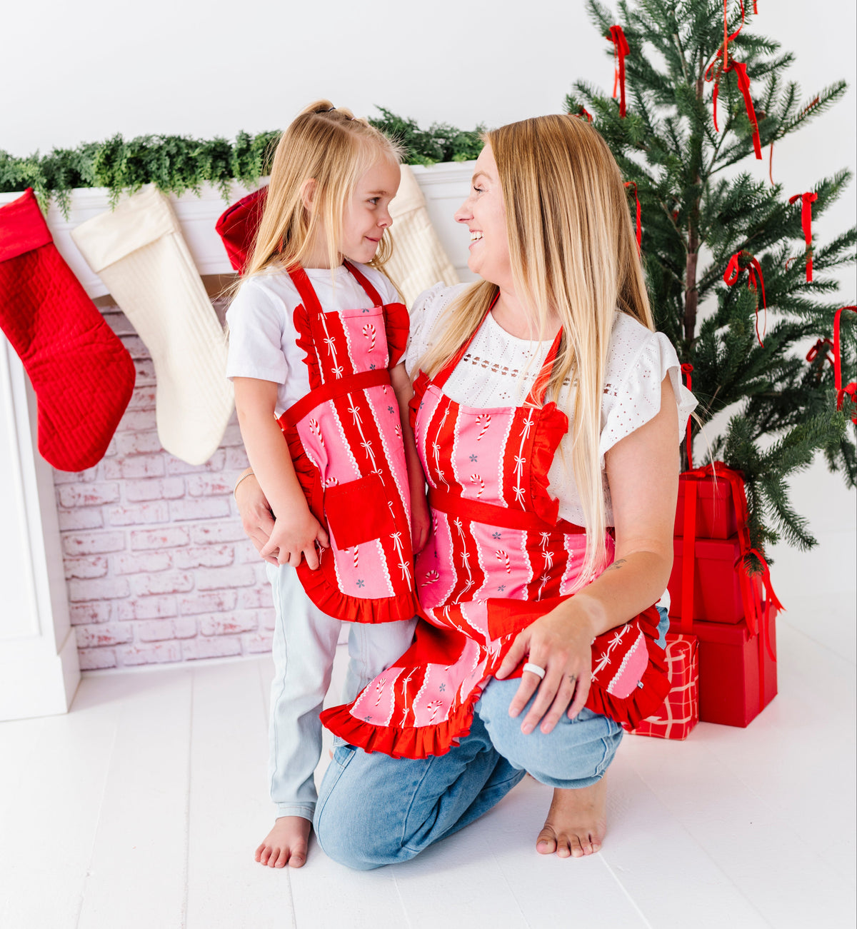 A woman and a young girl kneeling on a white floor in a decorated holiday room, wearing matching red and pink patterned aprons. A Christmas tree, stockings, and wrapped gifts are visible in the background.