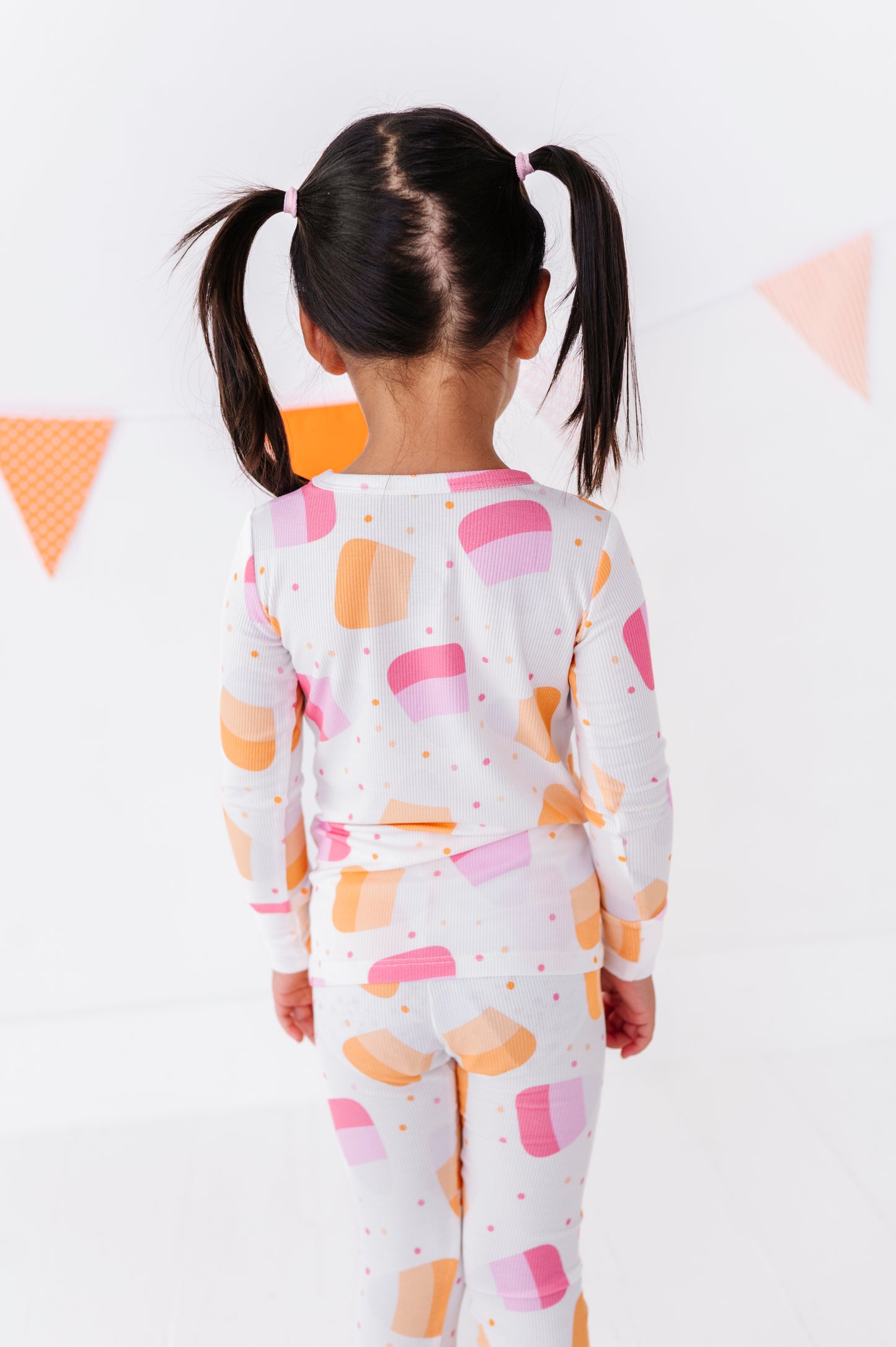 A young girl with dark hair in pigtails is shown from behind wearing a matching white long-sleeve top and leggings set with a colorful pattern of pink and orange semi-circles and small orange dots. The background is a clean white wall with a decorative orange and pink bunting banner.