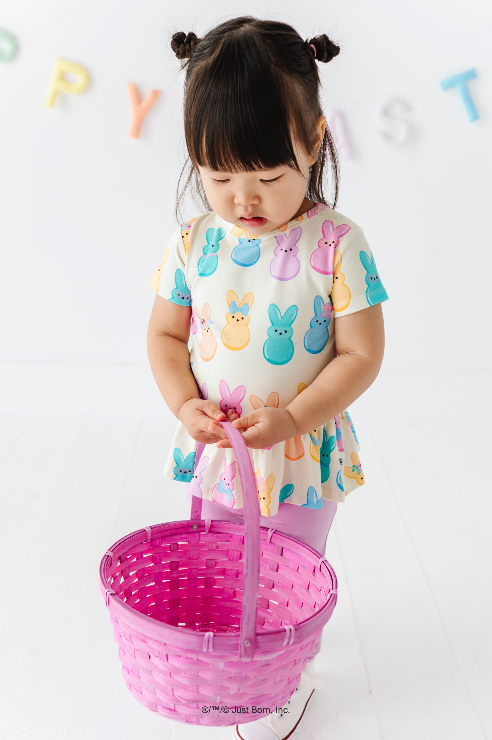 Child wearing a colorful shirt with PEEPS® bunny patterns, holding a pink basket against a white background.