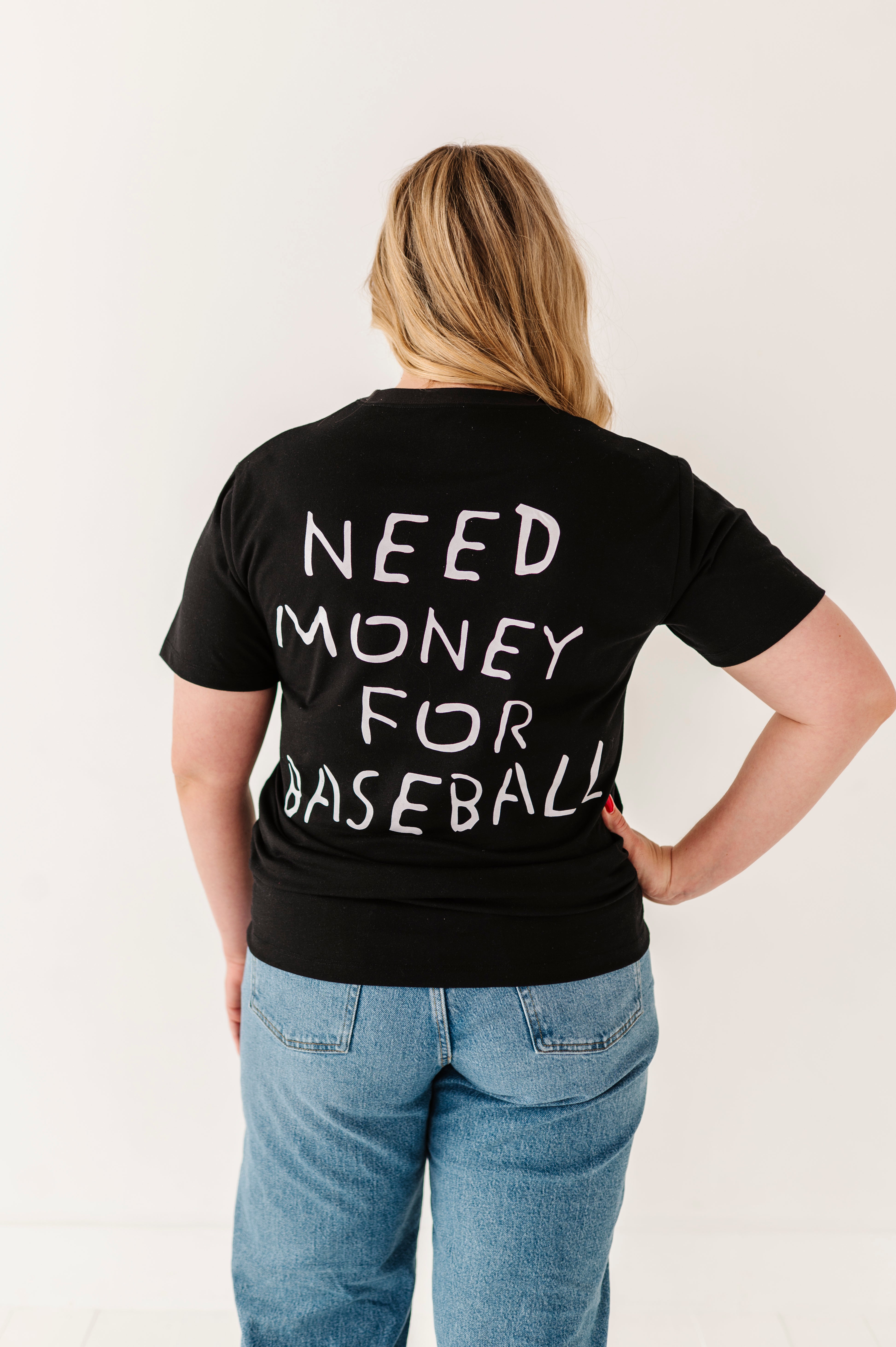 Person wearing a black t-shirt with 'NEED MONEY FOR BASEBALL' text on the back, standing against a plain background.