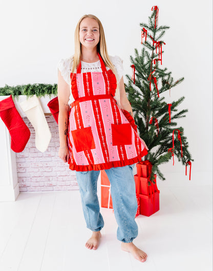 A woman stands barefoot in a bright white room wearing a red and white holiday-themed patchwork apron. To her left is a white brick fireplace with red and white stockings, and to her right is a small decorated Christmas tree with red ribbons and wrapped presents.