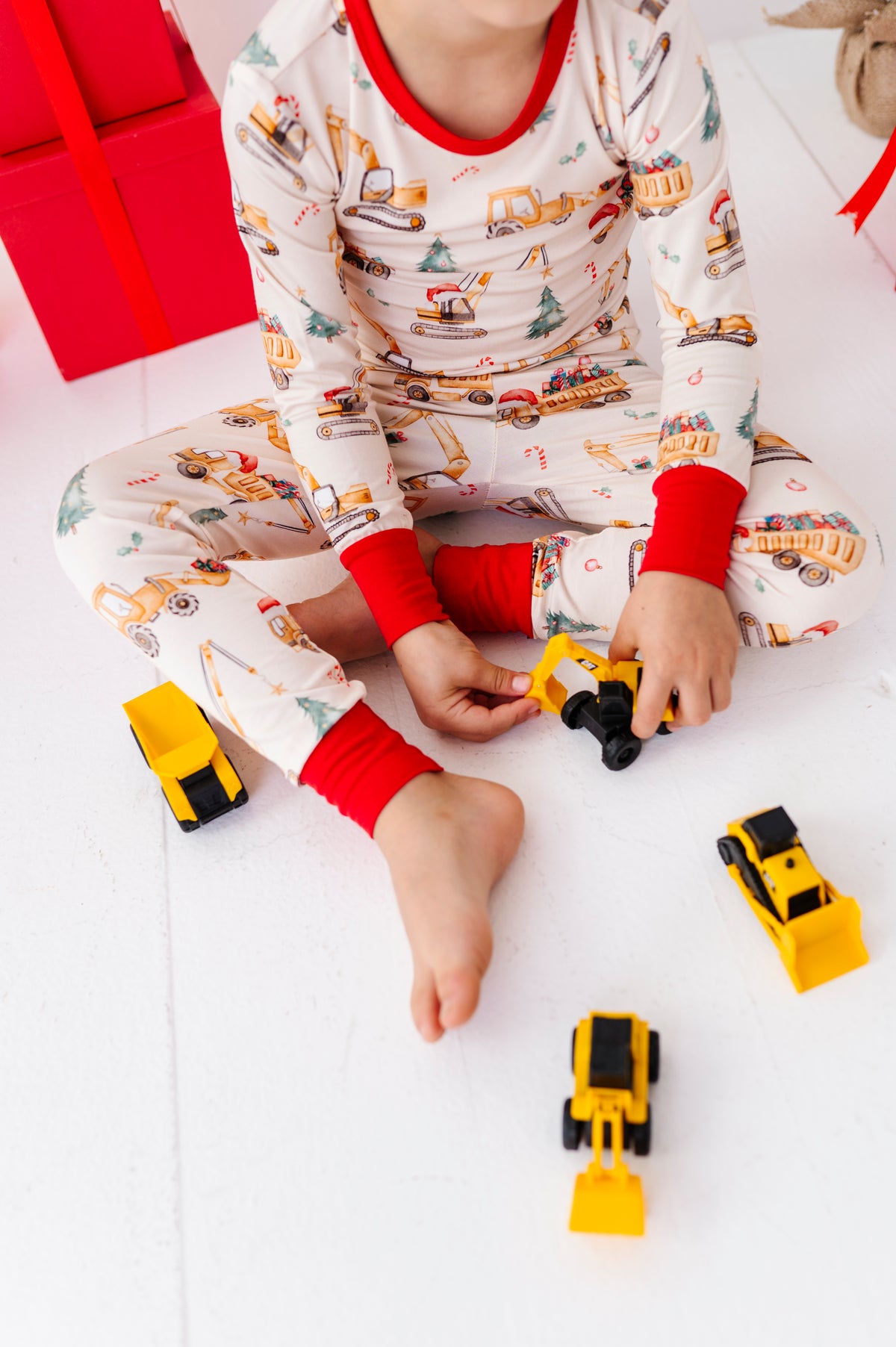A child sitting on a white floor wearing a cream bamboo pajama set with red cuffs and a construction vehicle print, holding a small yellow toy truck with other toy trucks around them.