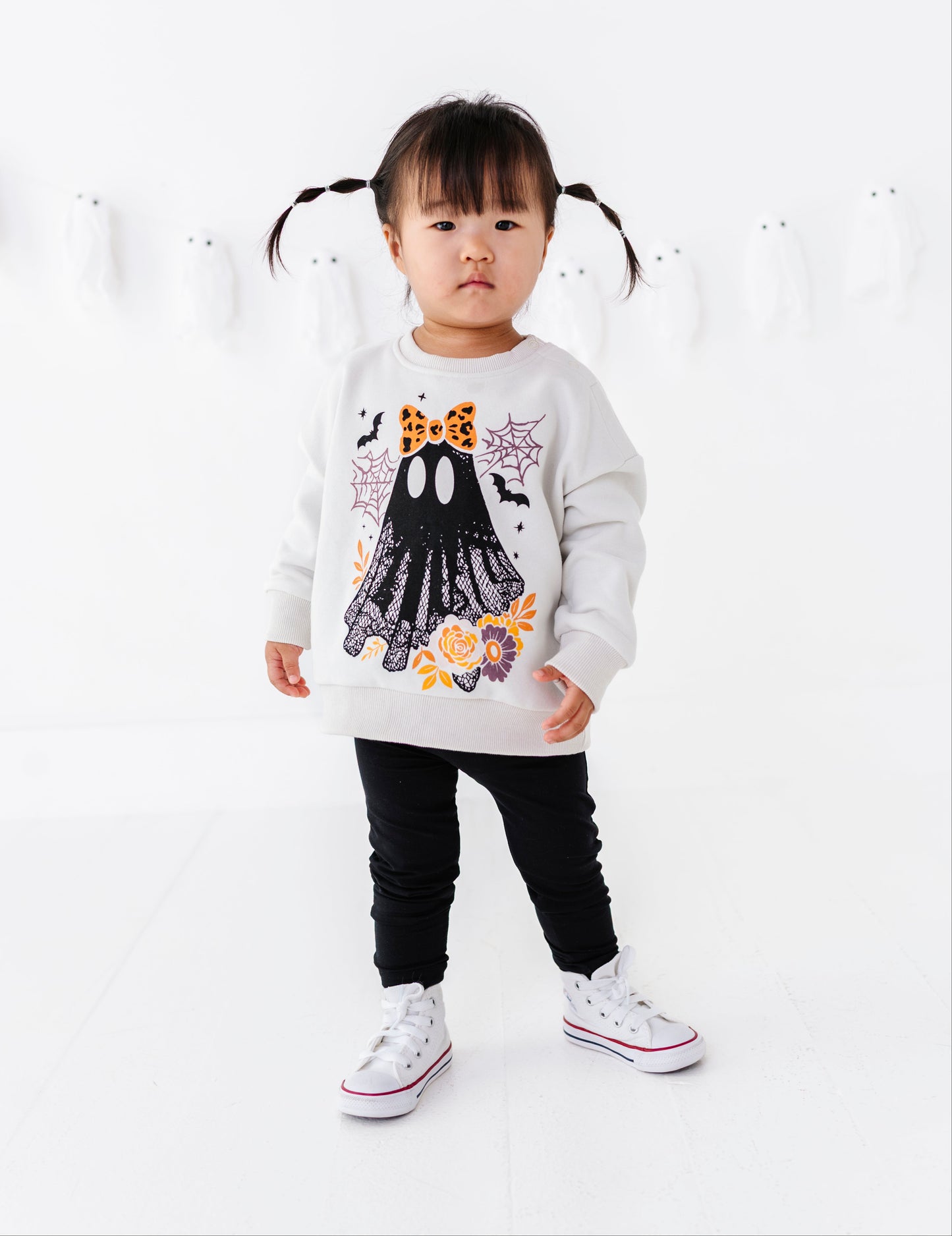 A toddler girl with black pigtails stands in a white studio wearing a white long-sleeve sweatshirt with a black ghost graphic and orange floral accents, paired with black pants and white sneakers.