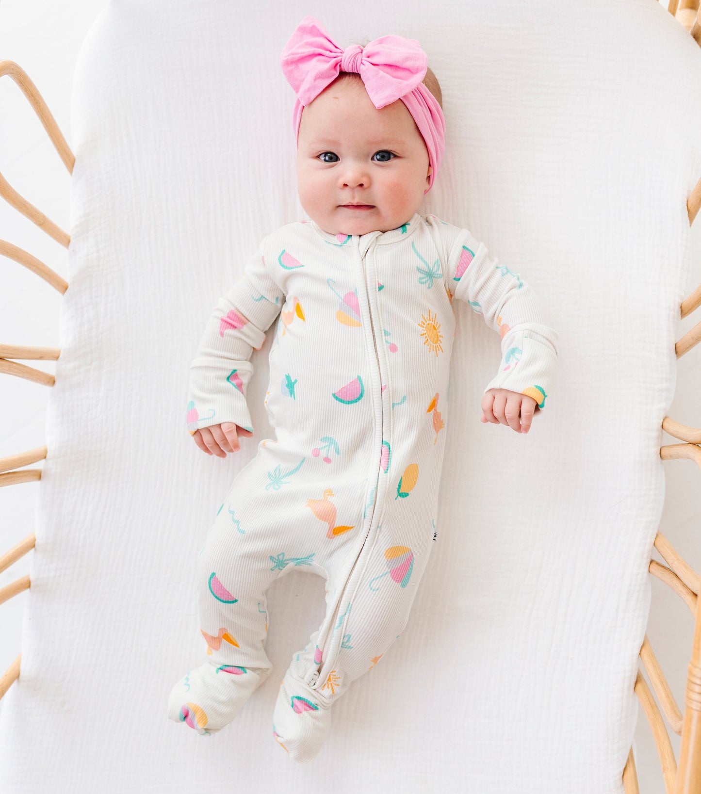 Baby in a colorful onesie with a pink bow sitting on a white cushion in a wicker chair.