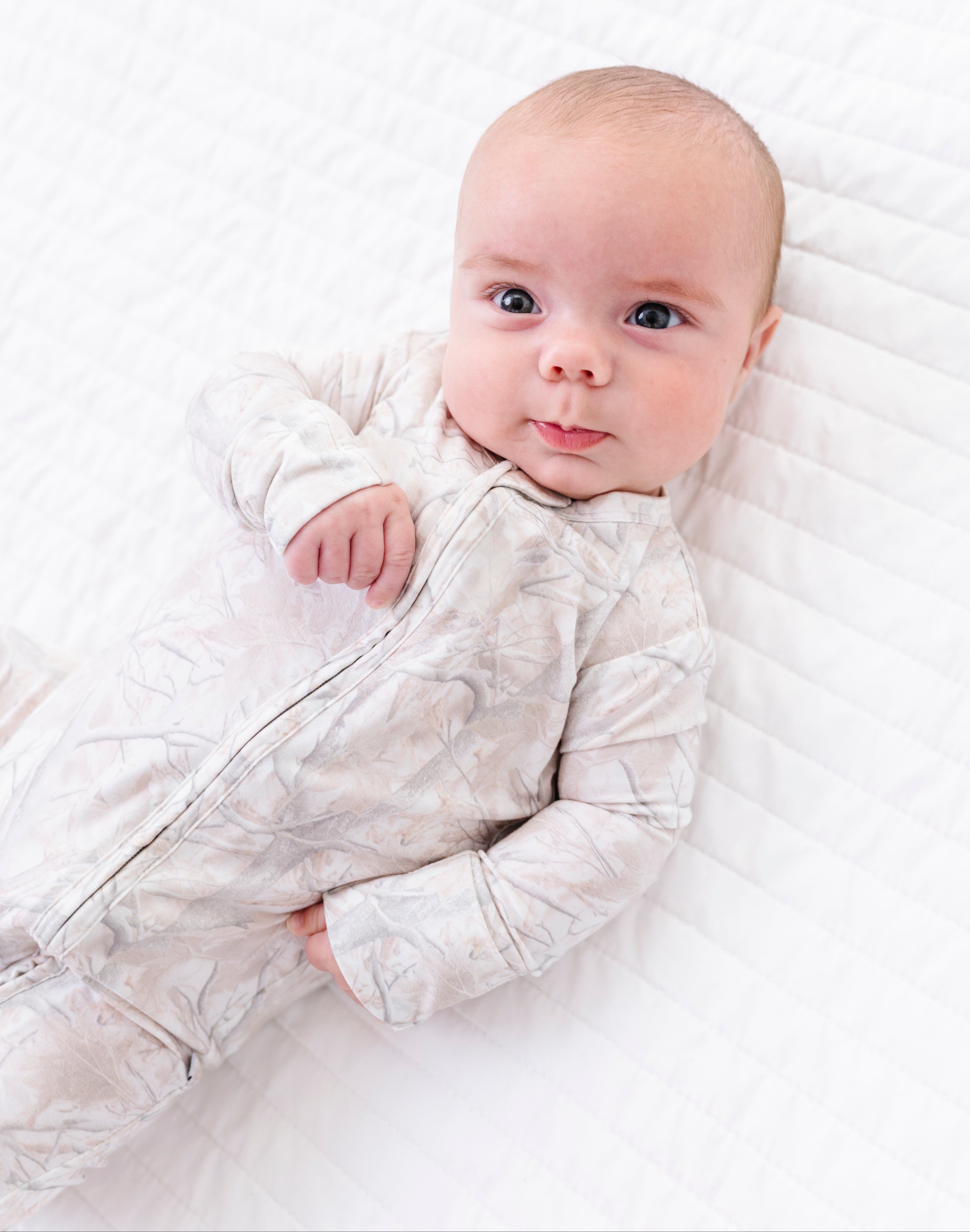 Baby lying on a white textured surface wearing a light gray camo onesie.