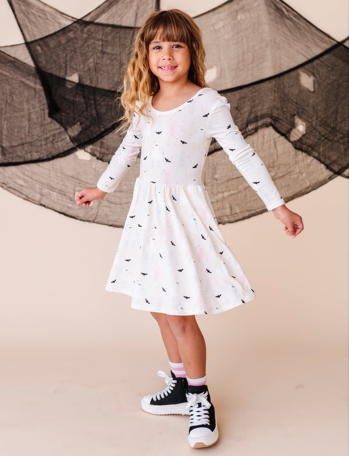 Young girl wearing a white dress with black patterns and black sneakers against a beige background
