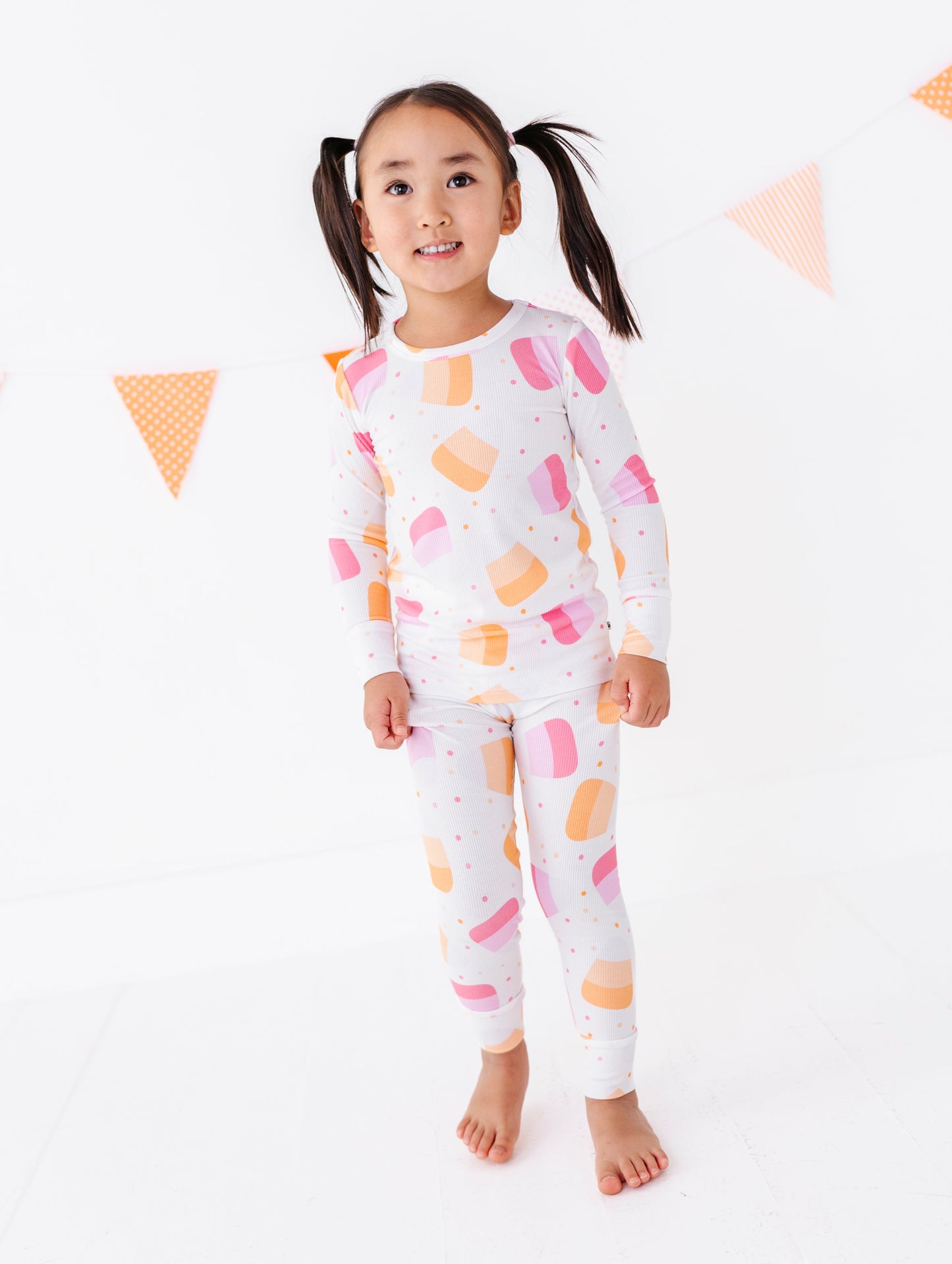 A young girl with dark pigtails stands barefoot against a white background, wearing a matching two-piece pajama set with a white base and a colorful ice cream cone pattern in pink, orange, and yellow.