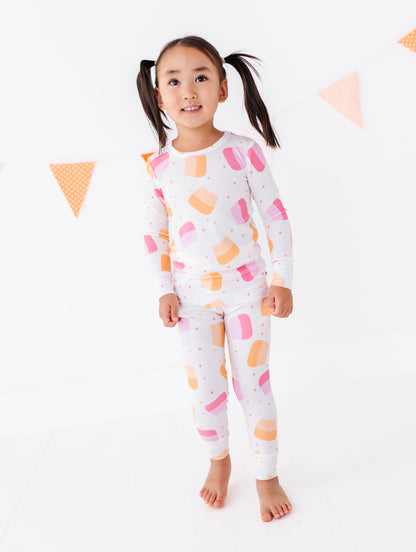 A young girl with dark pigtails stands barefoot against a white background, wearing a matching two-piece pajama set with a white base and a colorful ice cream cone pattern in pink, orange, and yellow.