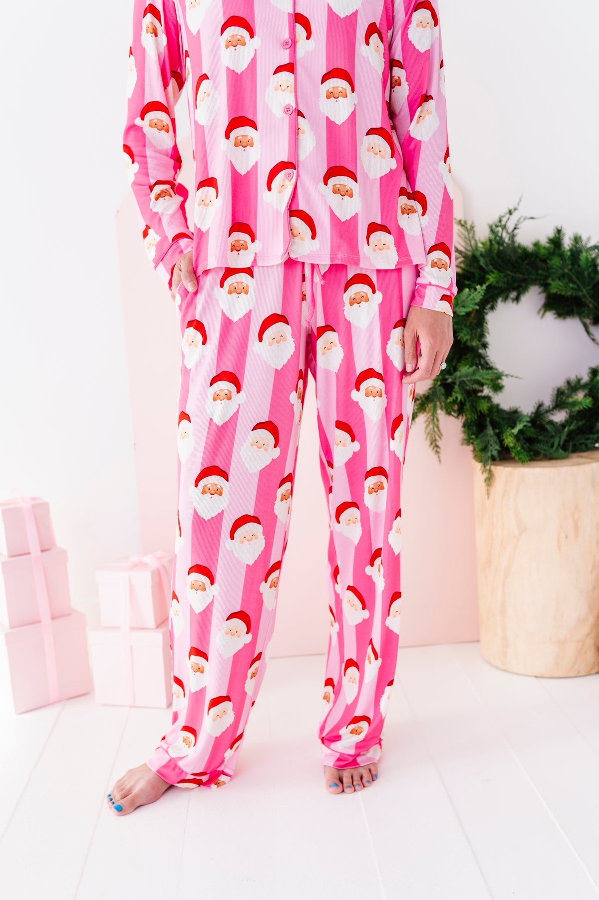 A close-up photo of a woman wearing pink Santa Claus bamboo pajamas, standing in a bright white room with a green wreath and wrapped gifts in the background.