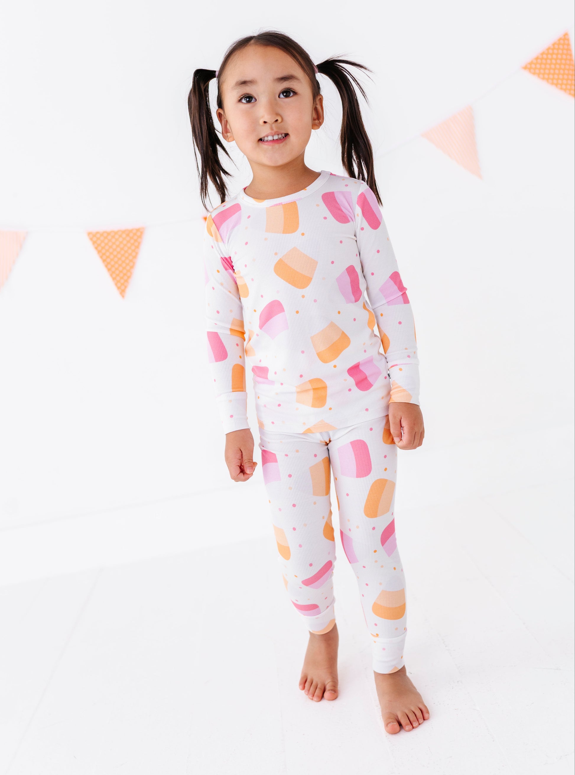A young girl with dark pigtails stands barefoot against a white studio background, wearing a matching long-sleeve pajama set with a colorful ice cream cone pattern in pink, orange, and yellow on a white base.
