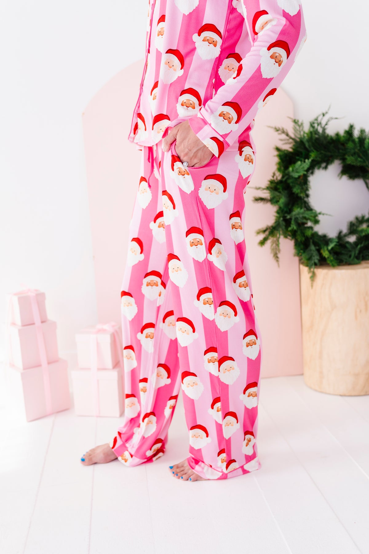 A close-up photo of a woman wearing pink Santa Claus bamboo pajamas, standing in a bright white room with a green wreath and wrapped gifts in the background.