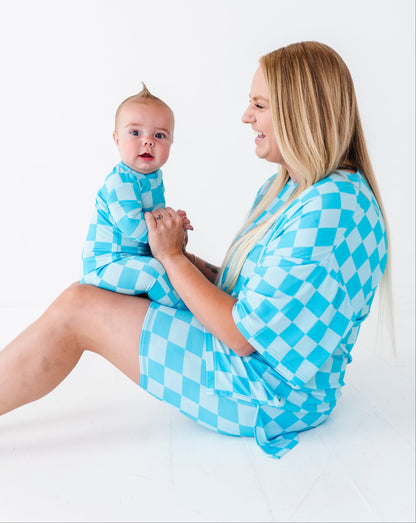 A woman and baby wearing matching light blue and white checkered outfits, sitting together on a clean white studio floor.
