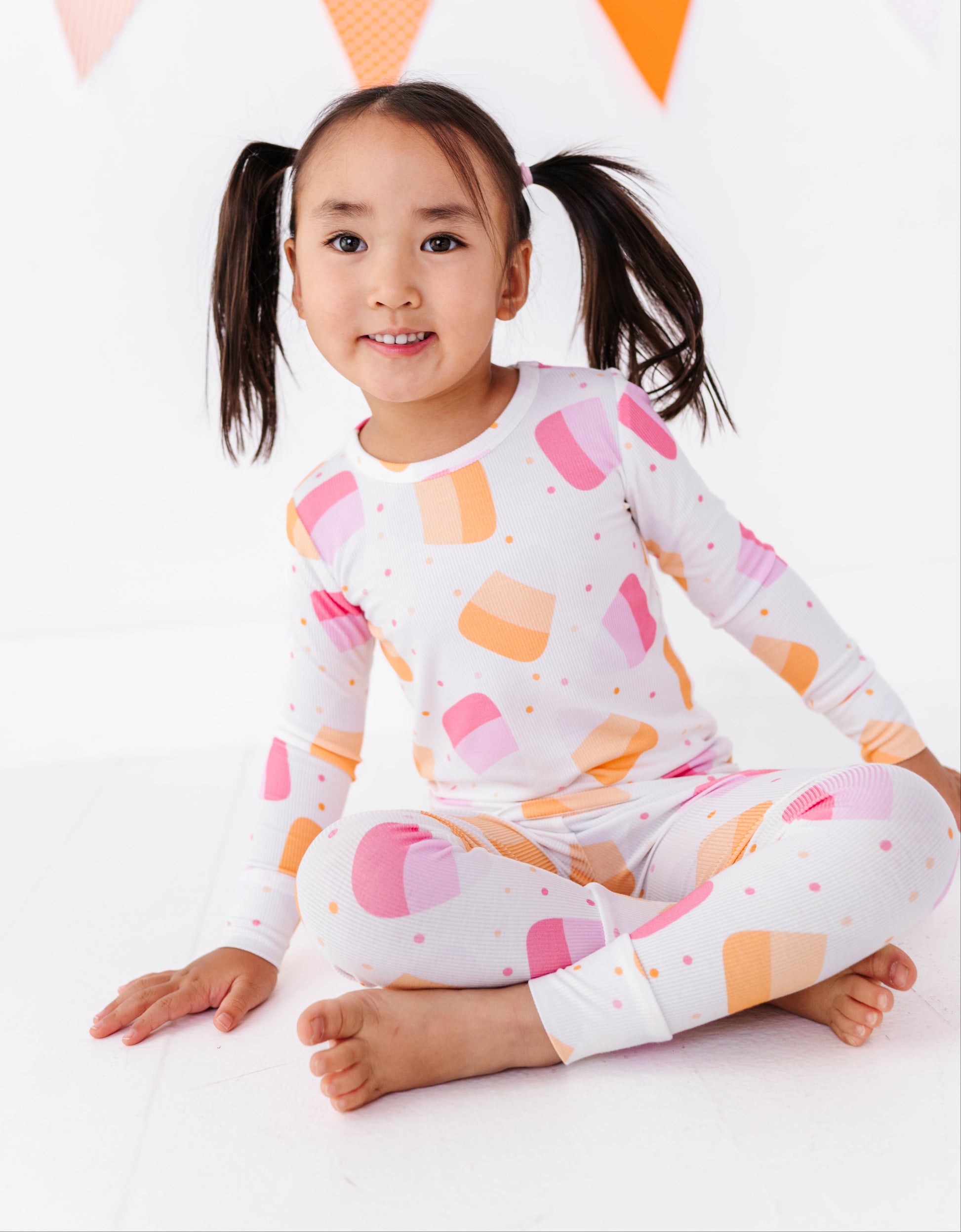 A young girl with dark hair in pigtails sits cross-legged on a white floor, wearing a white long-sleeve pajama top and matching leggings with a colorful pastel pattern of pink, orange, and peach ice cream cones. A string of orange and pink bunting is visible in the background.