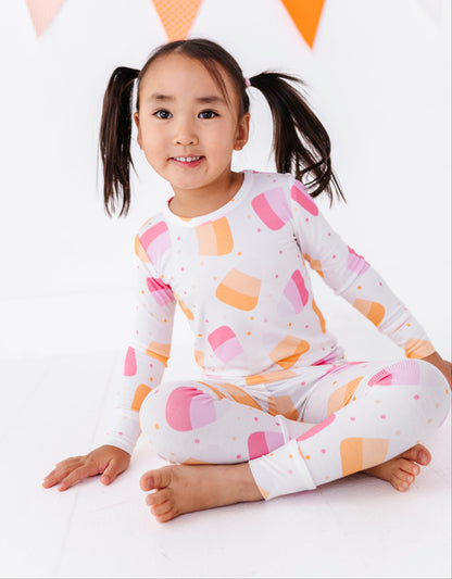 A young girl with dark hair in pigtails sits cross-legged on a white floor, wearing a white long-sleeve pajama top and matching leggings with a colorful pastel pattern of pink, orange, and peach ice cream cones. A string of orange and pink bunting is visible in the background.