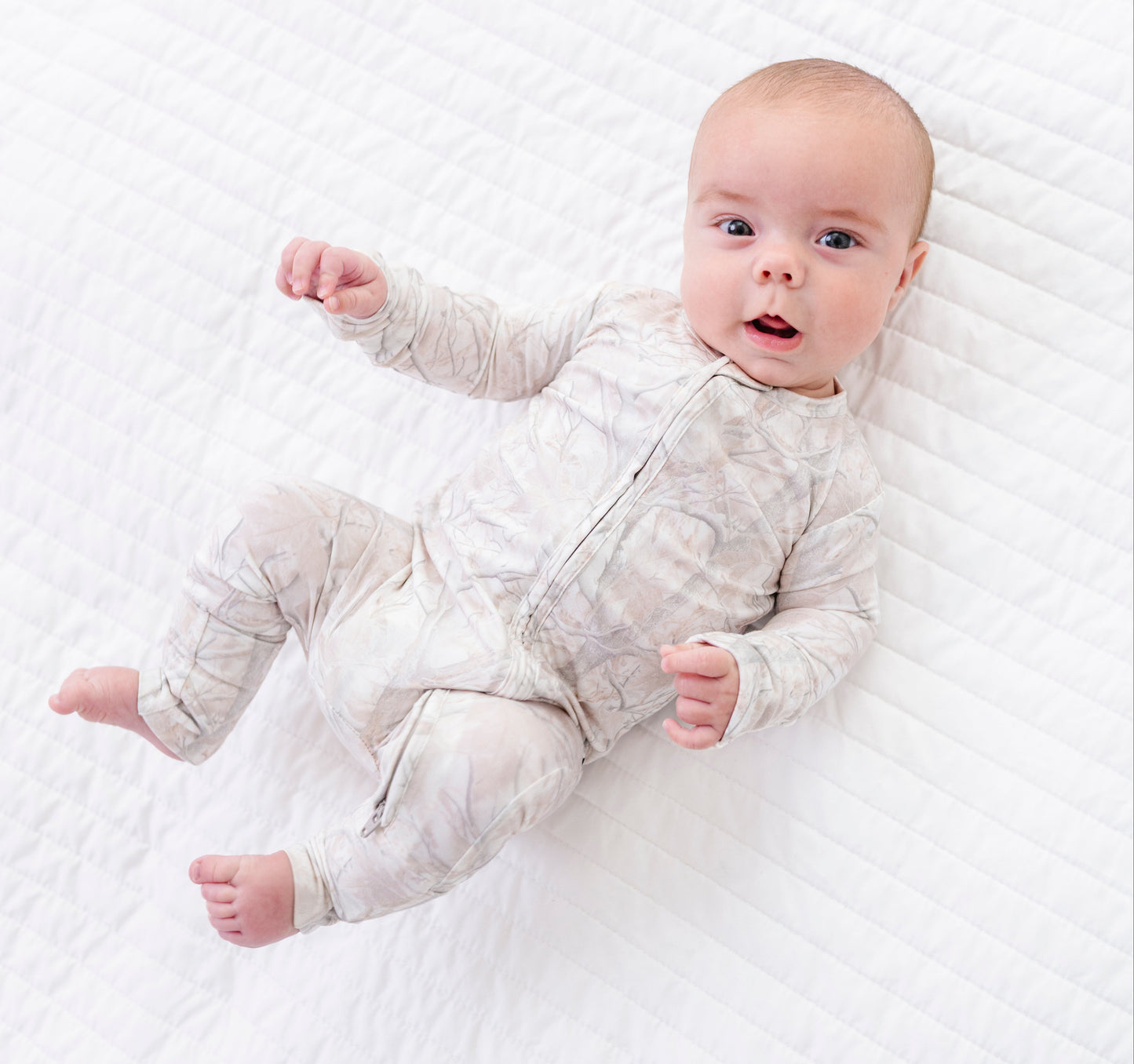 Baby lying on a white textured blanket wearing a light gray tree camo  romper .