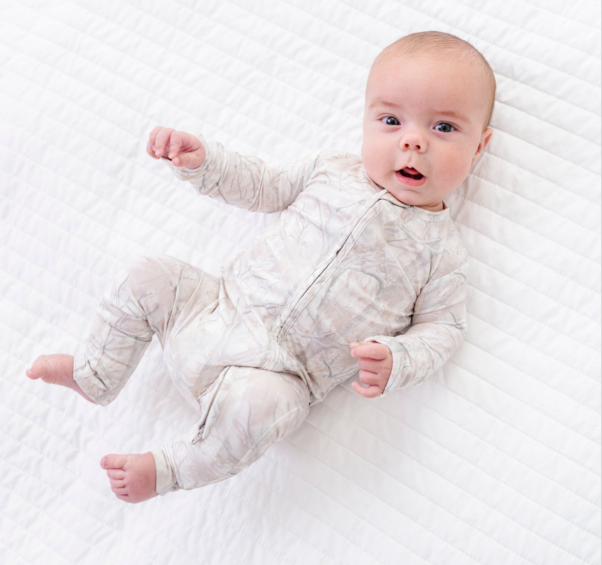Baby lying on a white textured blanket wearing a light gray tree camo  romper .