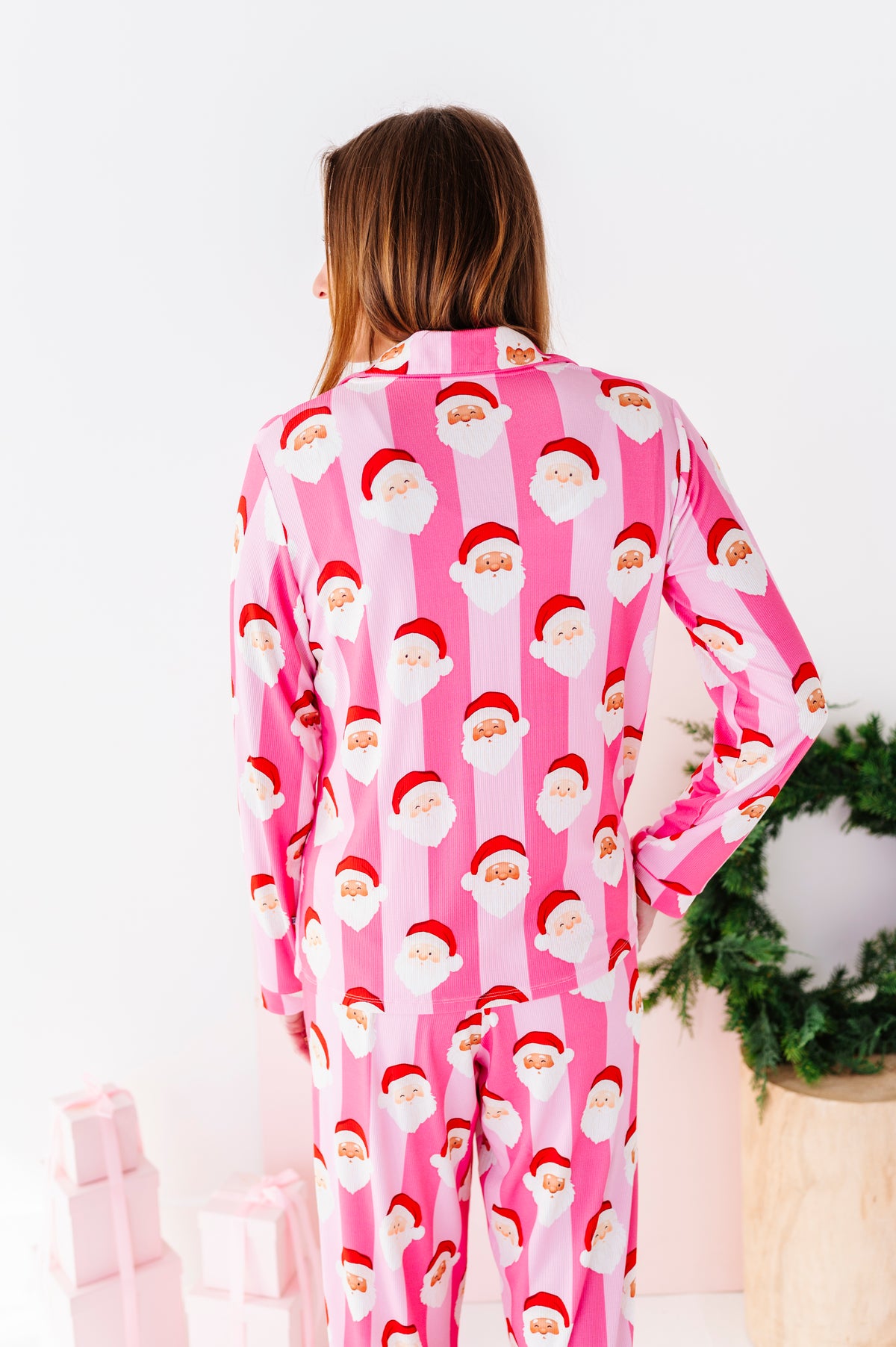A woman wearing pink Santa Claus bamboo pajamas, standing in a bright white room with a green wreath and wrapped gifts in the background.