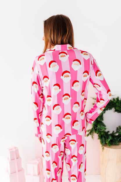 A woman wearing pink Santa Claus bamboo pajamas, standing in a bright white room with a green wreath and wrapped gifts in the background.