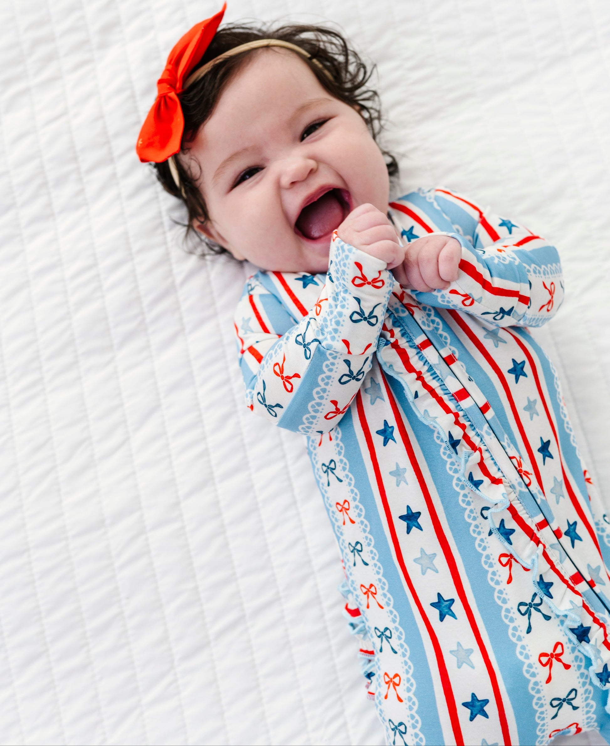 Baby wearing patriotic bamboo sleepwear with bows and stars, lying on a white textured surface.