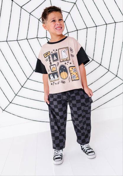 A young boy modeling a short-sleeve graphic t-shirt with black sleeves, paired with black and gray checkered pants and black-and-white sneakers, standing in front of a white background with a large black spiderweb graphic.