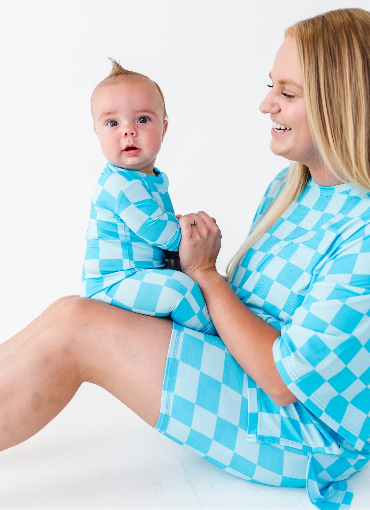 A woman and baby wearing matching light blue and white checkered pajamas, sitting on a white studio floor against a white background.