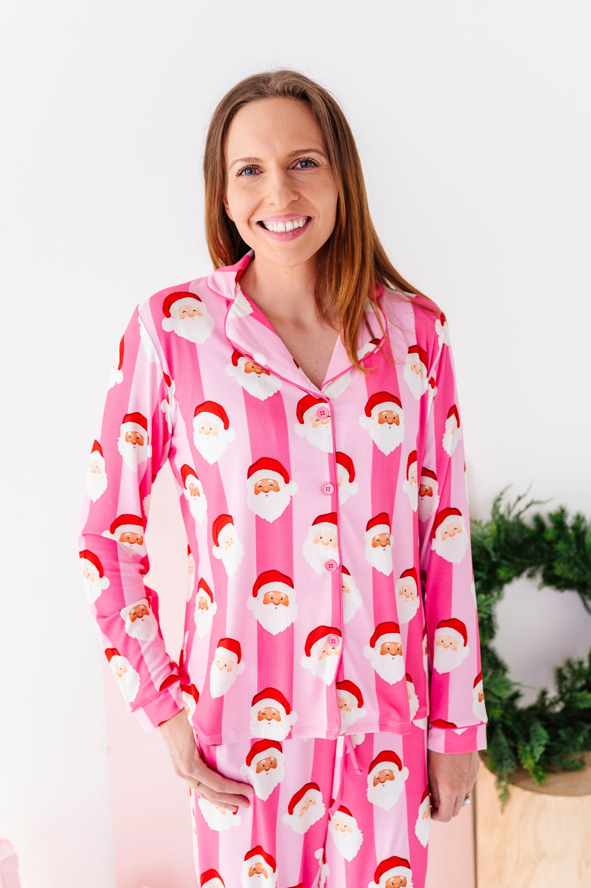 A smiling woman wearing pink Santa Claus bamboo pajamas, standing in a bright white room with a green wreath and wrapped gifts in the background.
