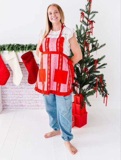 A woman stands barefoot in a bright white room wearing a red and pink patchwork apron over a white top and blue jeans. To her left is a decorated Christmas tree with red ribbons and wrapped presents, and to her right is a fireplace with red and white stockings hanging from a green garland.
